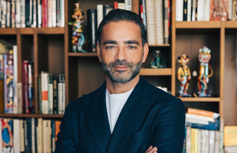 yiannis misirlis wearing a dark blazer over a white shirt stands confidently in front of a bookshelf filled with books and colorful decorative items.