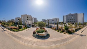 A gated residential complex with modern apartment buildings, landscaped gardens, and the "Sunset" entrance sign, under a bright blue sky.