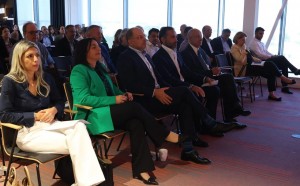 A group of real estate professionals seated attentively during a conference, with a modern, bright room featuring large windows in the background. The attendees, dressed in formal and business-casual attire, focus on the speaker, reflecting engagement in the event's proceedings.