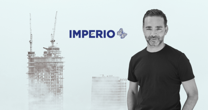 Yiannis Misirlis in a black t-shirt stands confidently in front of a hazy cityscape with a high-rise building under construction. The Imperio logo with a butterfly is prominently displayed next to him.