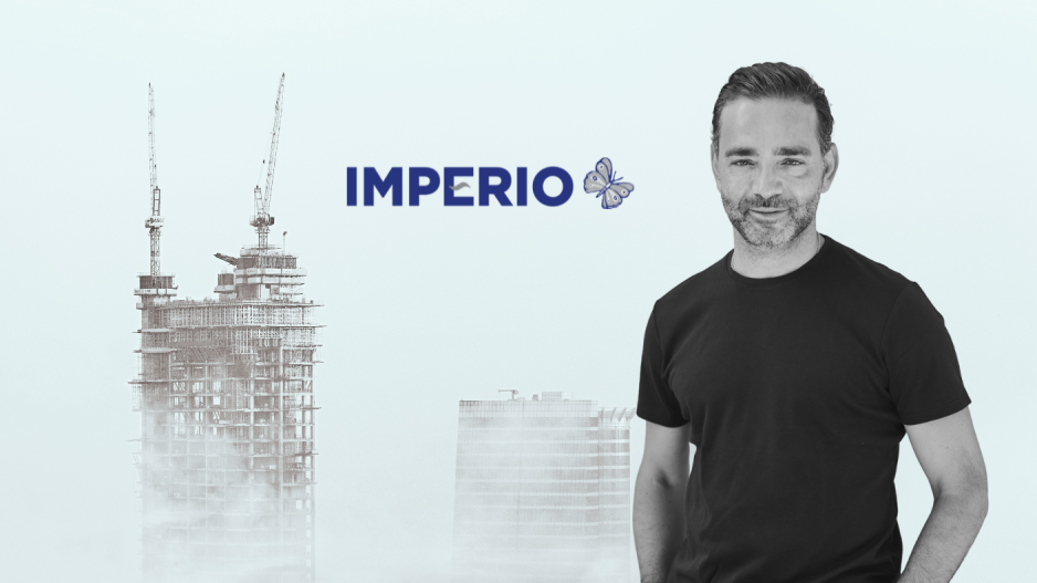 Yiannis Misirlis in a black t-shirt stands confidently in front of a hazy cityscape with a high-rise building under construction. The Imperio logo with a butterfly is prominently displayed next to him.