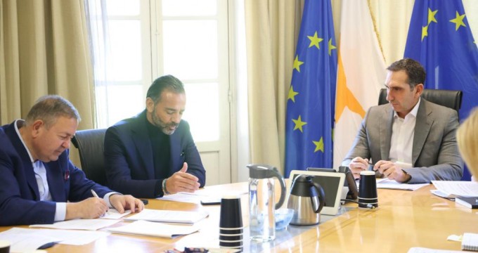 Three men in a formal meeting, seated around a wooden conference table with documents, cups, and a tablet, with the Cyprus and European Union flags in the background.