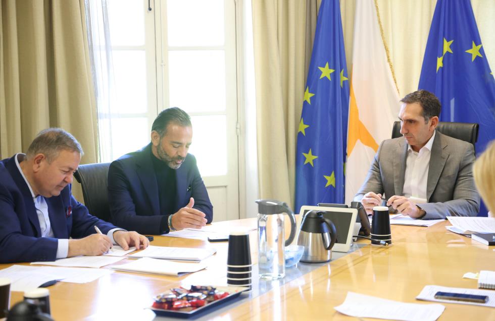Three men in a formal meeting, seated around a wooden conference table with documents, cups, and a tablet, with the Cyprus and European Union flags in the background.