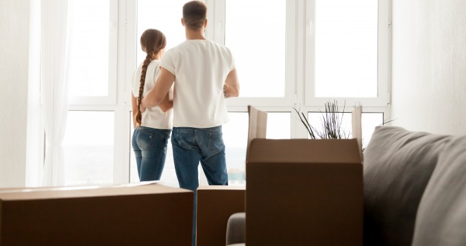 Millennial couple stand near window looking far away after moving in to new home with cardboard boxes, husband and wife plan future together, hugging seeing new perspectives. New beginning concept