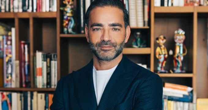 yiannis misirlis wearing a dark blazer over a white shirt stands confidently in front of a bookshelf filled with books and colorful decorative items.