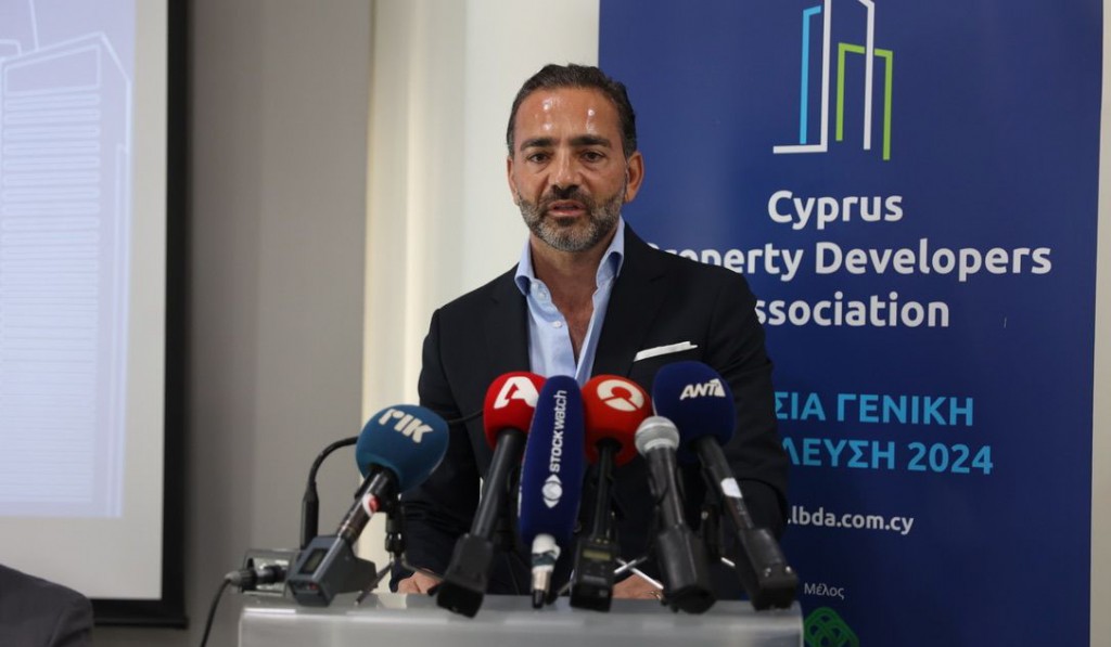 Yiannis Misirlis in a suit stands at a podium, speaking to multiple microphones from various media outlets, with a backdrop featuring the logo and name of the Cyprus Property Developers Association.