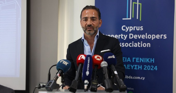 Yiannis Misirlis in a suit stands at a podium, speaking to multiple microphones from various media outlets, with a backdrop featuring the logo and name of the Cyprus Property Developers Association.